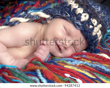 Sleeping newborn baby boy one week old with blue and white hat. He is laying on a blanket of multicolored yarn.