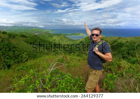 Young happy man standing with a backpack on a hill with a raised hand and looking at the camera