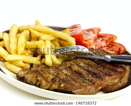 Delicious beef steak dinner with french fries tomatoes and utensils on plate against white background.