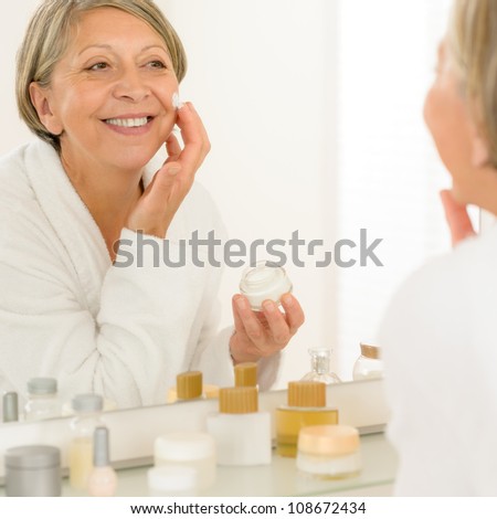 Smiling senior woman apply anti-wrinkles cream looking in bathroom mirror
