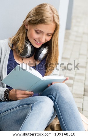 Smiling young student reading book outside school sitting on ground