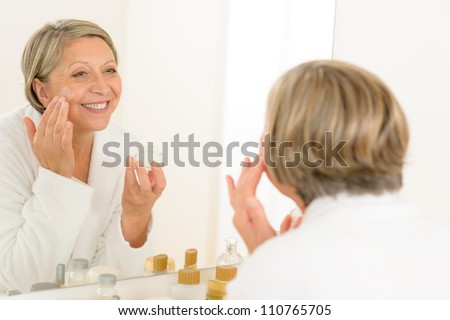 Senior woman looking in bathroom mirror and applying anti-wrinkles cream