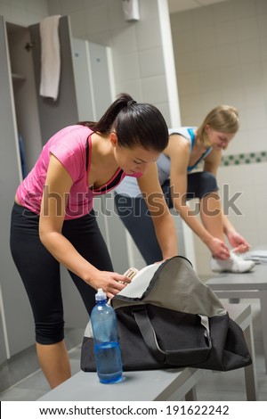 Woman packing bag at gym\'s locker room with friend in background