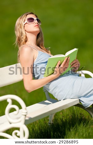 Young blond woman sitting on white bench in spring, enjoying sun and holding book; shallow DOF