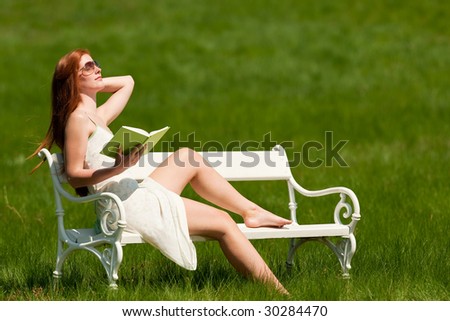 Red hair woman reading book on white bench in green meadow in spring