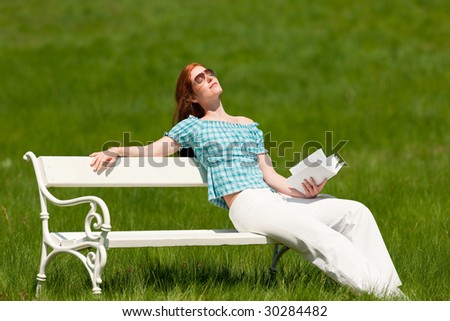Red hair woman with book enjoying sun on white bench in green meadow in spring