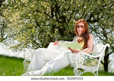 Red hair woman reading book on white bench in green meadow in spring