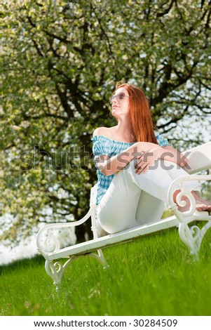 Red hair woman sitting on white bench in green meadow and enjoying sun in spring