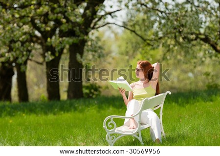 Red hair woman reading green book on white bench in a meadow in spring