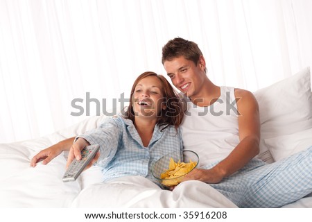 Young man and woman lying down in white bed watching television and eating crisps