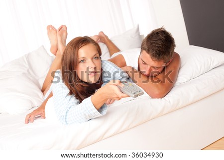 Young man and woman lying down in white lounge watching television together
