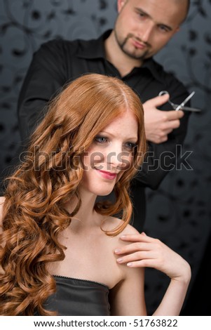 Professional hairdresser with long red curly hair fashion model at black luxury salon, hair cut with scissors
