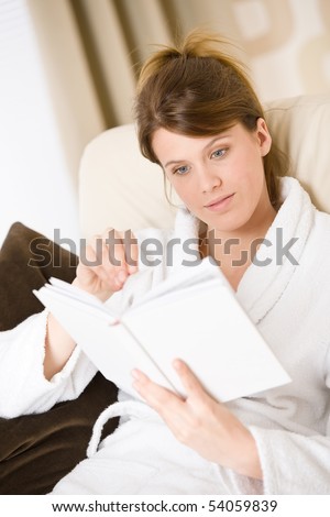 Young woman read book sitting on sofa relaxing in lounge wearing bathrobe