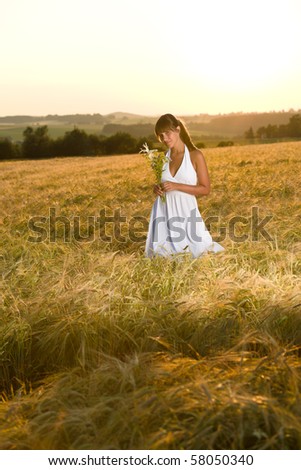 Romantic brunette woman in sunset corn field wear white dress, holding bouquet of flowers