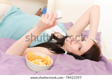 Young happy girl lying on bed eat chips hold phone