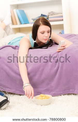 Young happy student girl on bed eat chips hold phone