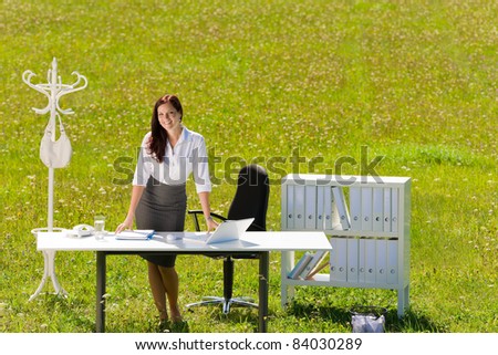 Young businesswoman in sunny meadow office smiling standing behind table