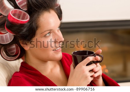 Woman with curlers holding mug sitting by fireplace red bathrobe
