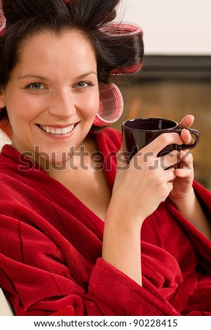 Woman with curlers holding mug sitting by fireplace red bathrobe
