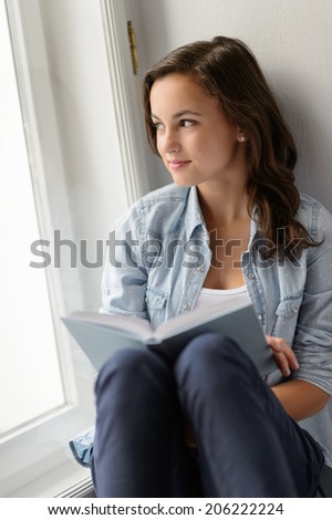 Teenage student girl sitting with book by window looking away daydreaming