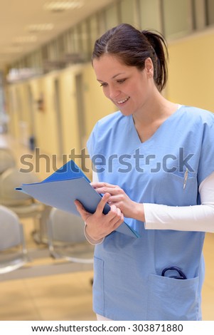 Smiling young female doctor in hospital looking down at patient records standing alone in hallway