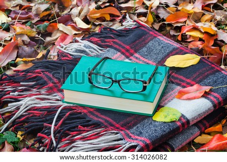 Cozy blanket, a book and glasses among fallen in the garden of autumn leaves . The items needed for a relaxing holiday in the autumn.