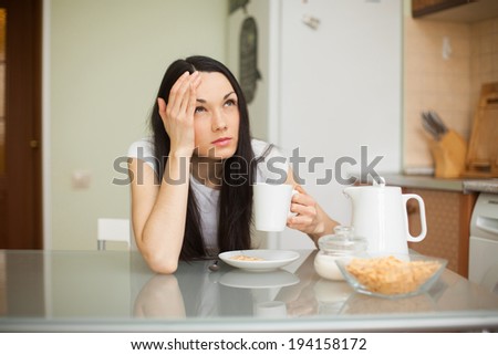 brunette girl having breakfast in the kitchen with headache holding cup of coffee