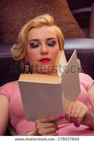 Relaxed casual blonde woman in pink cardigan holding a book lying on a dark brown couch