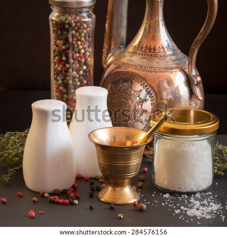 Concept of salt and pepper accessories. Mortar, jars with salt and pepper, old copper jar, dried thyme, porcelain salt and pepper on black stone background. Square