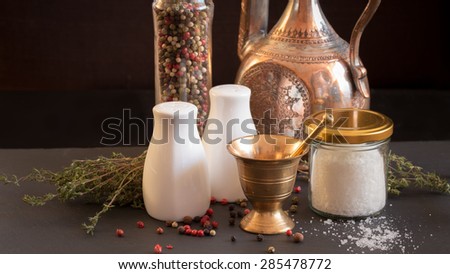 Concept of salt and pepper accessories. Mortar, jars with salt and pepper, old copper jar, dried thyme, porcelain salt and pepper on black stone background. Horizontal, wide screen format