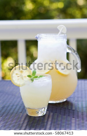 Glass cup of lemonade with ice, sprig of mint and crushed ice and pitcher behind with lemon slices and crushed ice inside.  Selective focus on the glass cup.