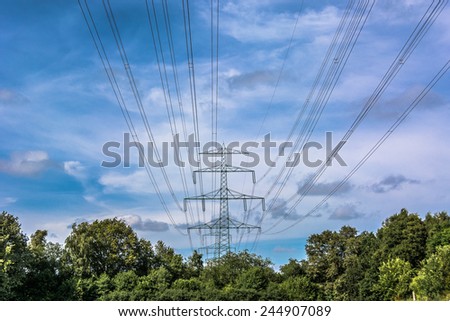 Electricity Pylon tower in landscape with green trees against sky background \
Blue and cloudy sky behind an Electricity Pylon tower in landscape with green trees