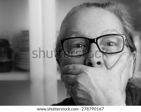 Amazed middle aged woman with hand in front of mouth \
Surprised senior woman with hipster style glasses covering her mouth. Black and white image
