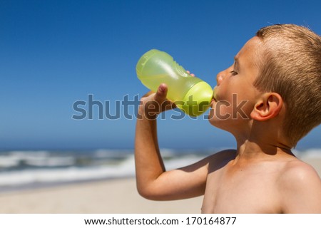 Upper body shot of thirsty boy who is drinking water from his bright green water bottle. Boy is standing at beach while the sun is shining without mercy. Ocean waves and blue sky in the background.