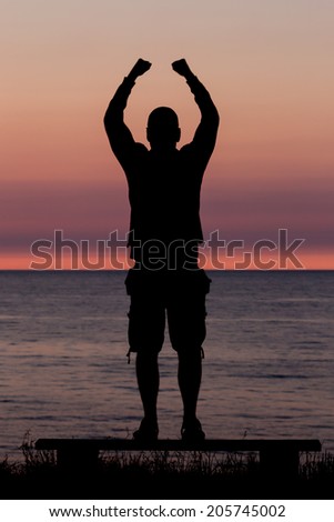 Silhouette of male person standing on a bench near the ocean at sunset and celebrating a major achievement.