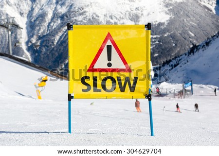 Yellow warning sign with the text slow and exclamation mark placed on ski piste at ski resort.