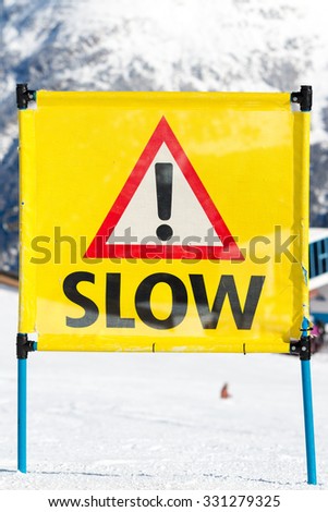 Yellow warning sign with the text slow and exclamation mark placed on ski run at ski resort.
