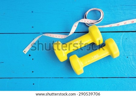 Yellow dumbbells and measuring tape plank on blue painted wood background.