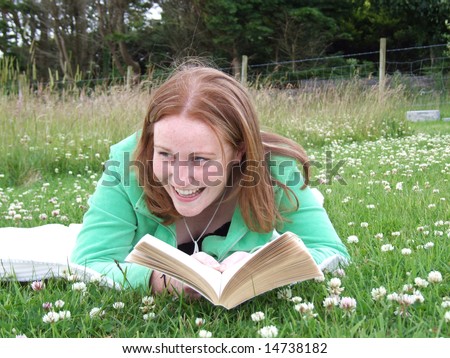 Pretty young girl with red hair laughing while reading a book laying on a blanket in meadow
