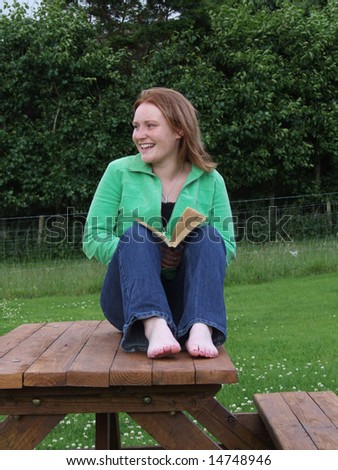 Pretty young girl with red hair sitting on a garden bench reading a book