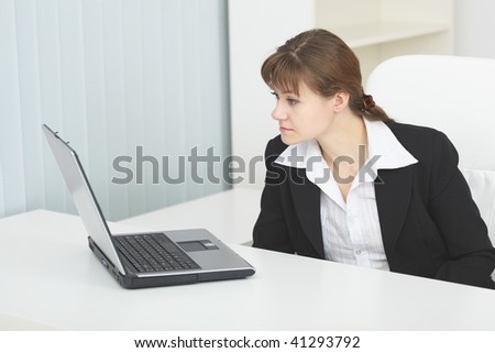 The young serious woman sits at a table at light office with the laptop