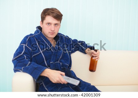 The guy sits on a sofa and drinks beer looking the TV