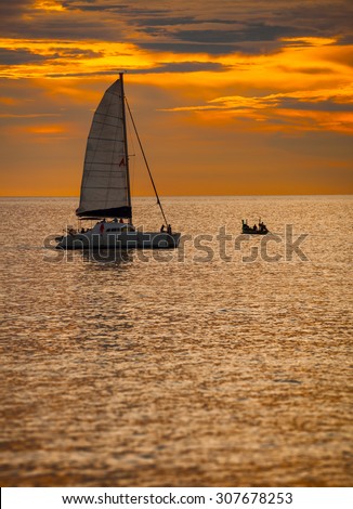 Sky blazing with the last orange light of sunset over a catamaran sailboat, with its sail unfurled, on a tropical sea.