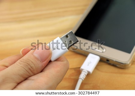 Hand holding USB connector, USB cable with smartphone on wood table.
