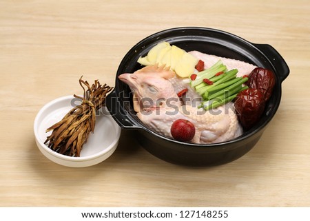 whole chicken prepared in a black china cooking pot with yellow ginger pieces, red chinese dates, red chinese hawthorn, green onion, red wolf berry and dried day lily for stewing on a wooden cut board