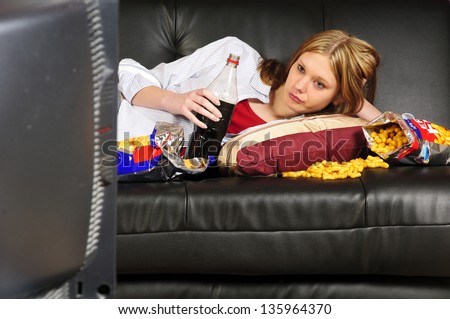 A Young Woman, Teenager With Long Blond Hair Lolls On A Black Leather Sofa, Watching Television And Eating Crisps And Coke Here.