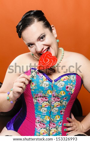 Attractive overweight woman sitting on a chaise longue and holding a big red heart shaped lollipop in hand. She wears a colorful corset. She grins at the camera and bites into the lollipop with joy.