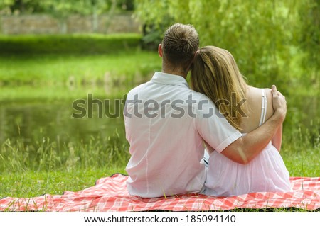 Young couple sitting together in a summer park and have a picnic on a checkered picnic blanket. You see them from behind as they hug and look at a lake.