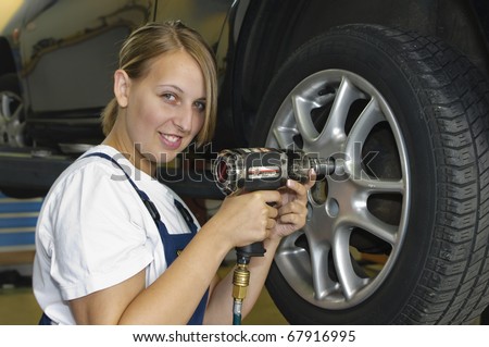 Female auto mechanic changing a wheel in a car in the garage with an impact wrench and smiling at the camera
