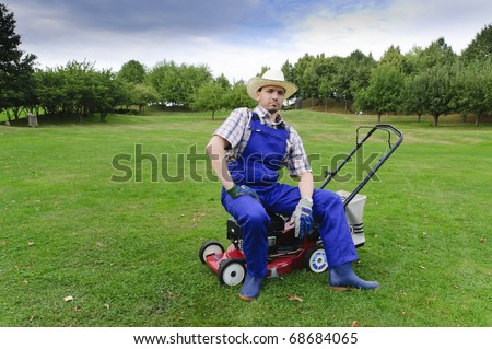 Adult man in blue (gardener), rubber boots and straw hat standing on a large park-like grounds, leaning proudly on his gas lawn mower.